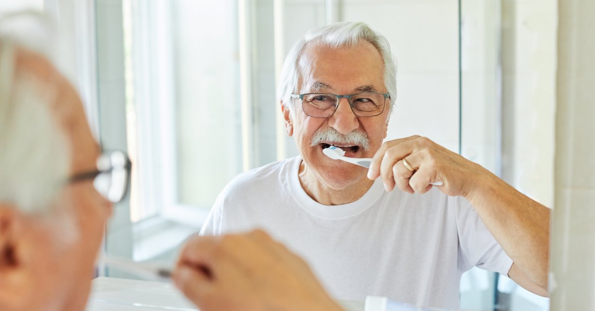 An older man in glasses and a white t-shirt standing in front of a mirror, looking at his reflection as he brushes his teeth.
