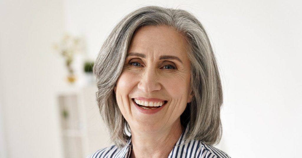 An older woman with a tidy gray bob haircut dressed in a striped shirt, smiling warmly in a room with white walls.