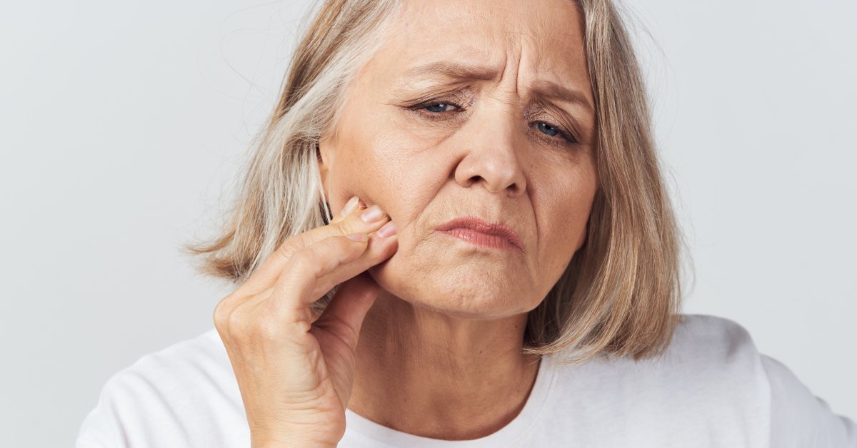 A woman in a white t-shirt pinches the skin on her lower cheek, looking uncomfortable and concerned.