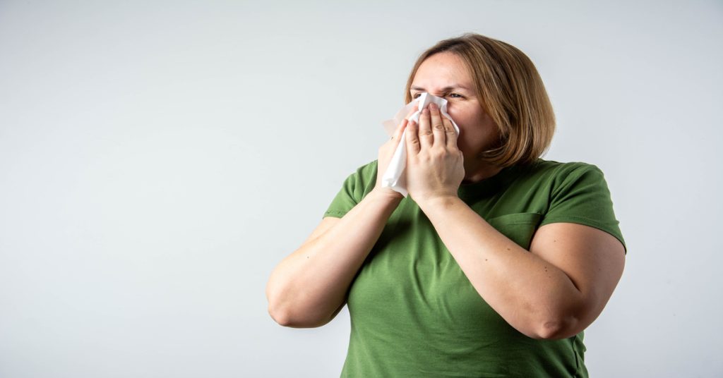 A woman wearing a green shirt scrunching her face as she holds a tissue to her nose with both hands.
