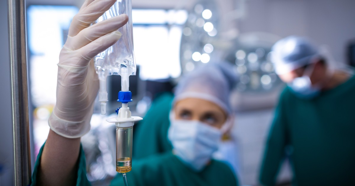 An individual in green scrubs, a mask, a hair net, and gloves inspecting an IV in a clean, white, sterile room.