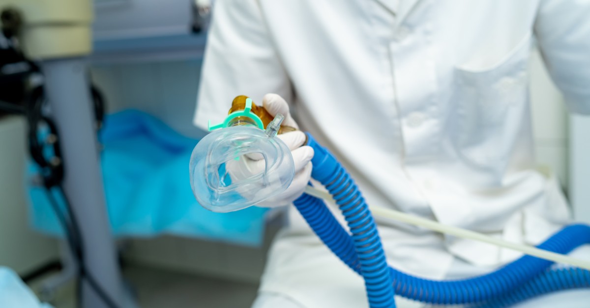 A person dressed in white scrubs sitting in a chair, holding a clear mask connected to a long blue tube.