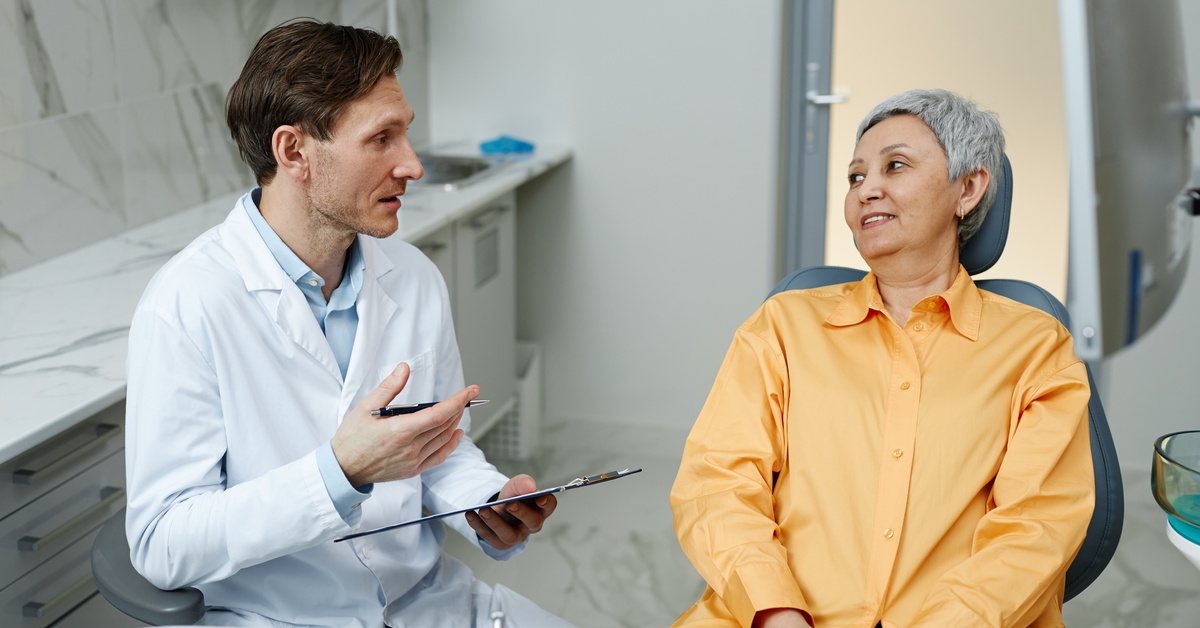 A man wearing a white coat and button-up shirt holding a clipboard while speaking to an older woman seated in a dental chair.