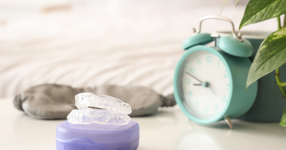 A bedside table with a sleep mask, alarm clock, potted plant, and a nightguard resting on its purple case.