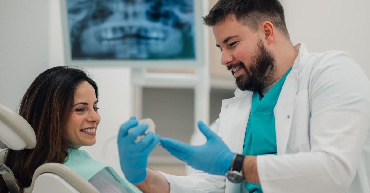A man in a white coat sitting next to a woman in a reclined dental chair, showing her a model of a tooth.