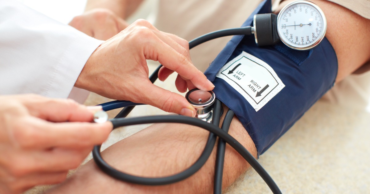 A person in a white lab coat attaching a blood pressure monitor to the upper left arm of another person in front of them.