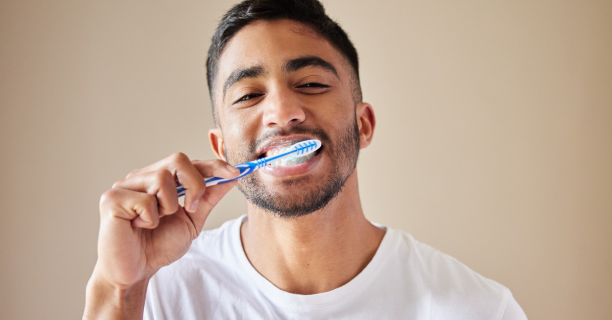 A man wearing a white T-shirt standing in a room with tan walls and using a blue toothbrush to brush his teeth.