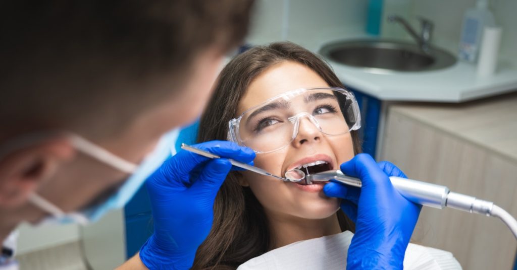 A man wearing a mask and gloves using a dentist's mirror and plugger to fill the tooth of a woman on an exam table.