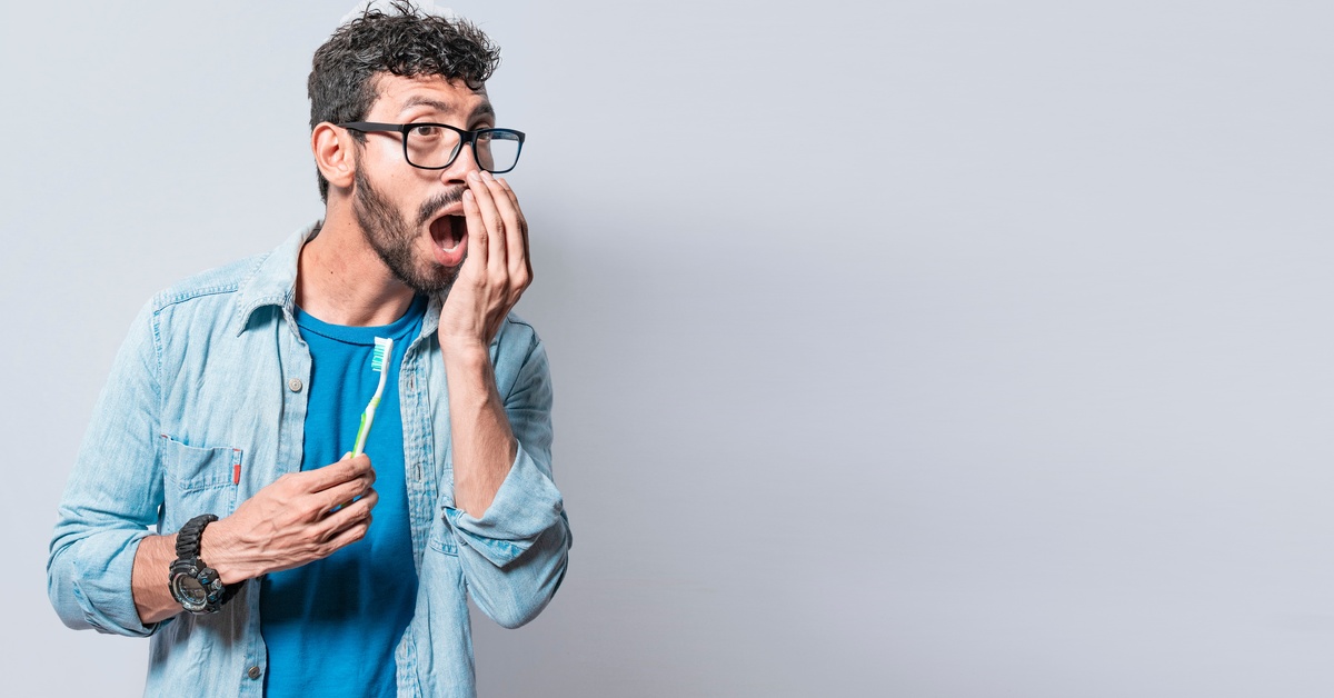 A man holding a toothbrush in one hand. He holds his other hand up to his mouth as he checks his breath.