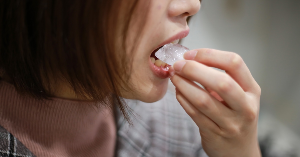 A close-up of a woman holding and eating a piece of ice with her mouth open and her hand near her face.