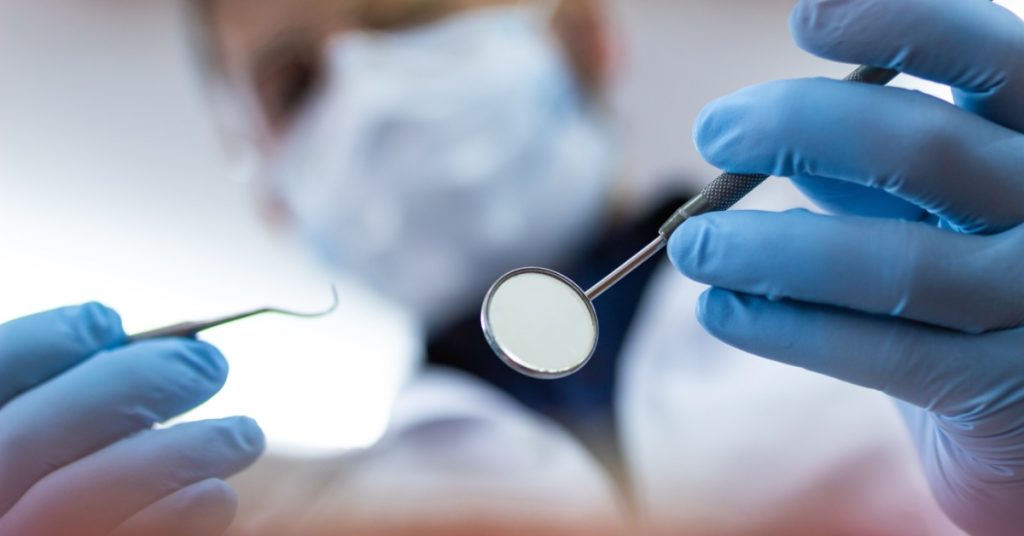 A blurry view from a patient perspective showing a dentist holding a mouth mirror and hook tool in their gloved hands.