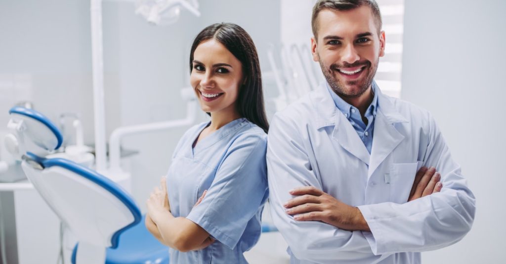 Two dental professionals wearing medical uniforms and coats standing side by side with their arms crossed.