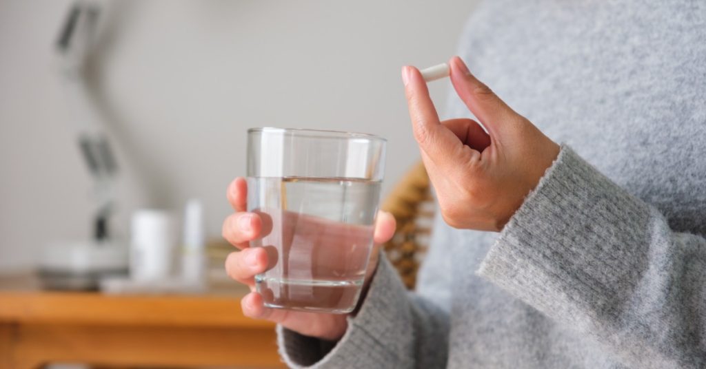 A woman wearing a gray turtleneck sweater holding white pills in one hand and a glass of water in the other.