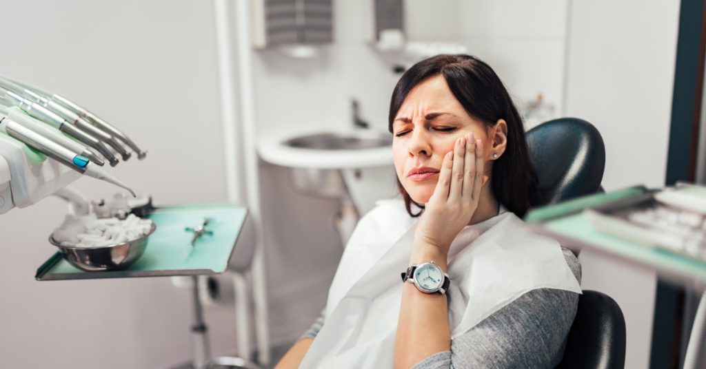 A woman sitting in a examination chair in a dental office, holding a hand up to her cheek while grimacing.