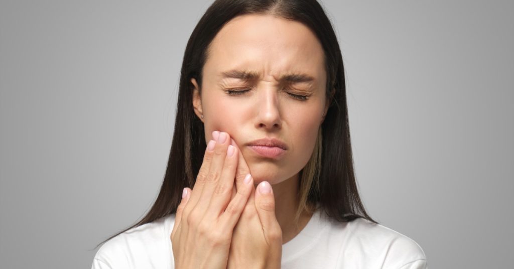 A woman wearing a plain white T-shirt pressing her hand to her cheek with eyes closed while experiencing tooth pain.