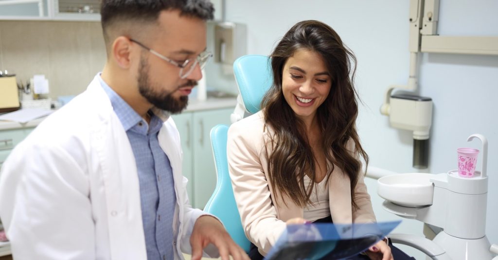 A patient sitting in a dental chair while a dentist explains a teeth x-ray on a monitor in a clinic.