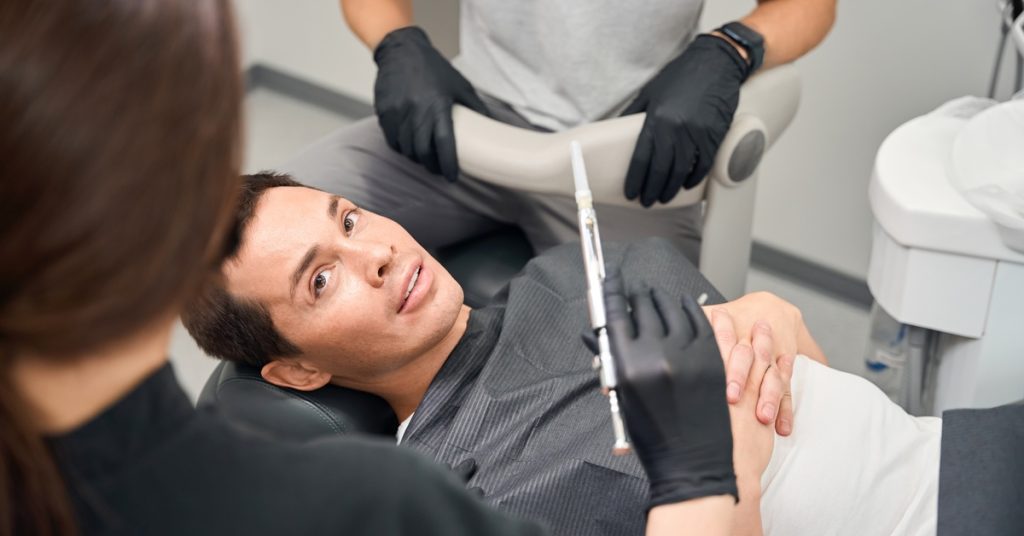 A woman wearing black gloves holding a dental syringe near a patient in a chair inside a dental office.