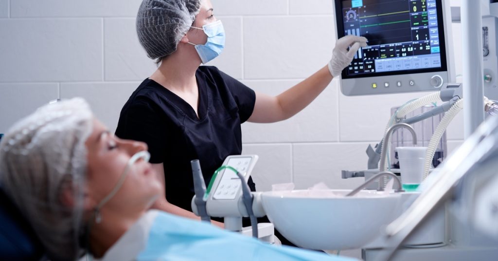 A woman wearing medical scrubs looking at a patient monitor beside a reclined person in a dental office.