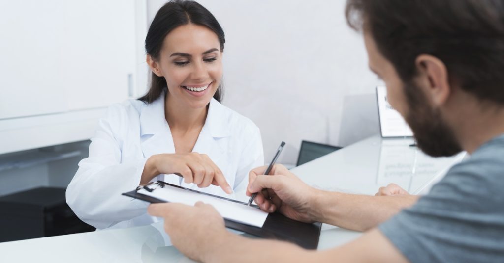 A person at a medical reception desk reviewing paperwork with another person seated across the counter.