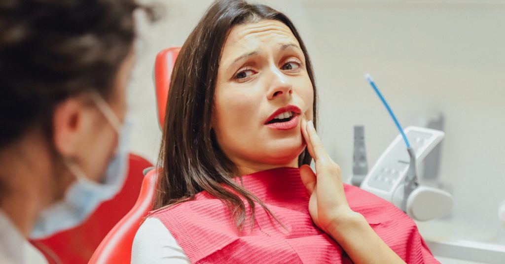 A young girl sitting in a dental chair while holding her cheek, with a medical professional nearby in an exam room.