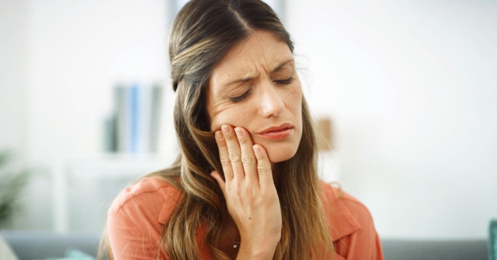 A woman pressing her hand to her cheek with closed eyes while sitting indoors in a coral shirt against a softly blurred room.