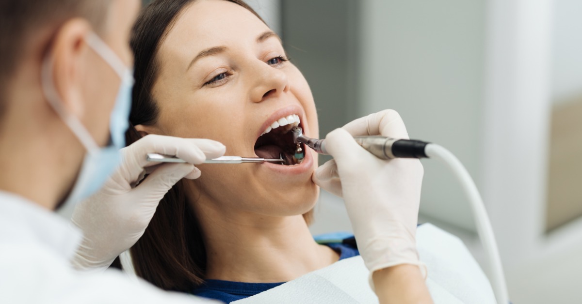 A person reclining in a dental chair while gloved hands use metal tools inside their open mouth during care.