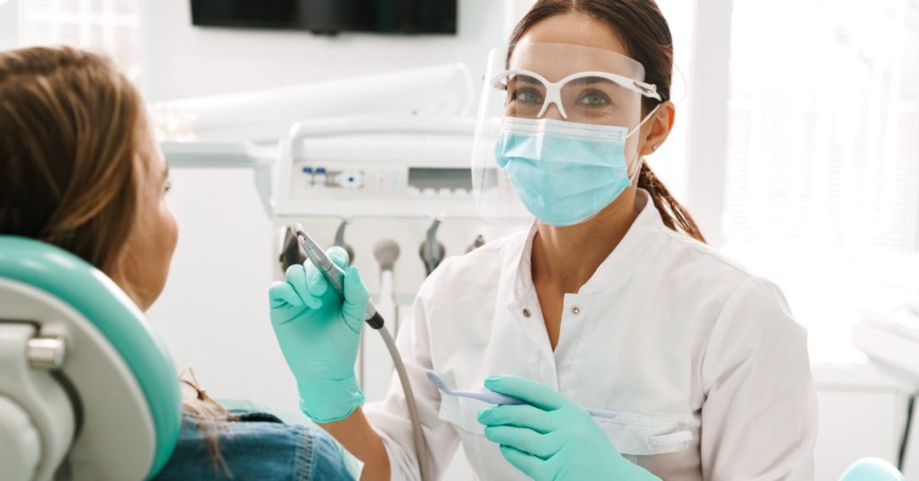 A masked woman in protective glasses holding dental tools beside a reclining person in a bright dental office.