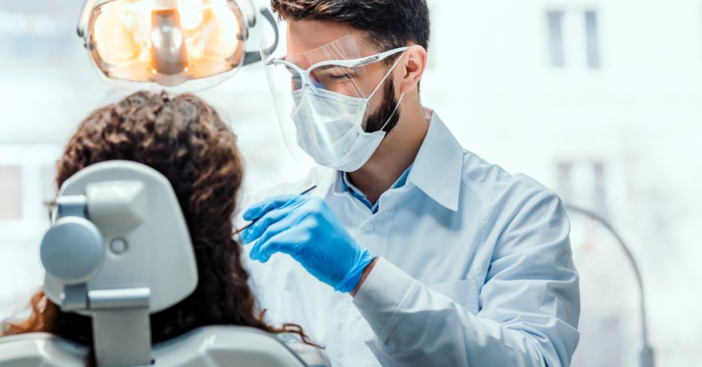 A person in protective eyewear and a mask examining a seated patient beneath an overhead dental light in an office.