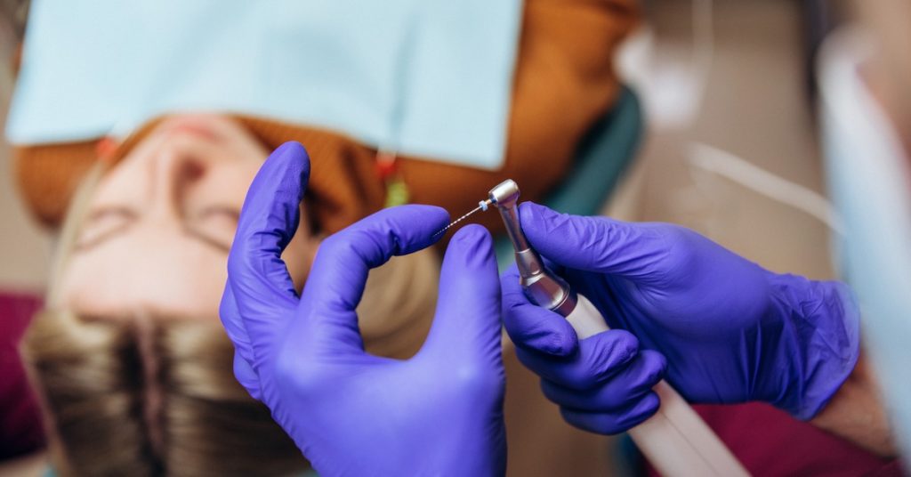 A gloved person holding a dental handpiece and small tool near a reclined patient in a dental chair.