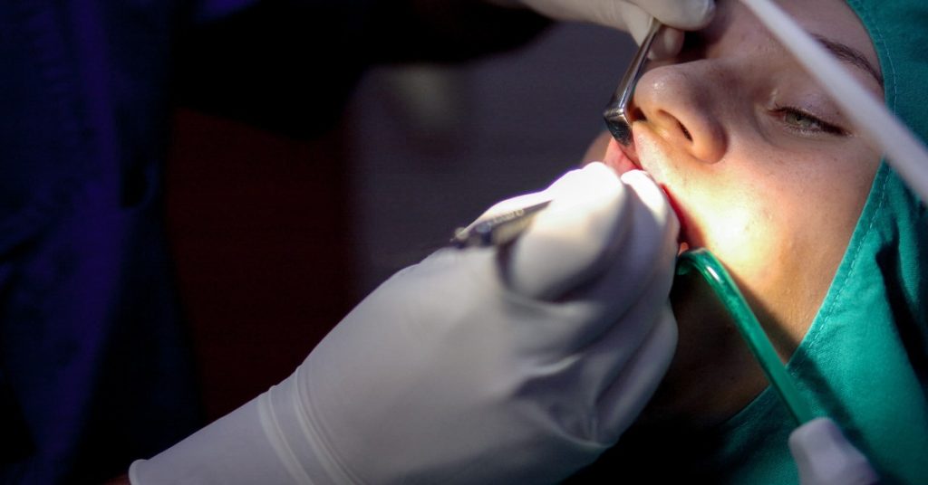 A person wearing green surgical fabric receiving dental work with gloved hands and metal tools near their mouth.