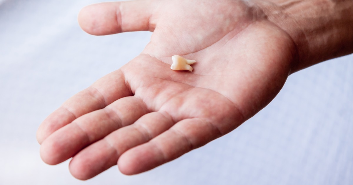 An open hand holding a small tooth with visible roots over a pale blue background, with fingers stretched outward.
