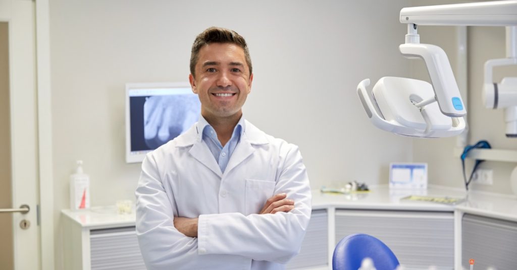 A smiling man in a white coat standing with crossed arms in a dental office with exam equipment nearby.