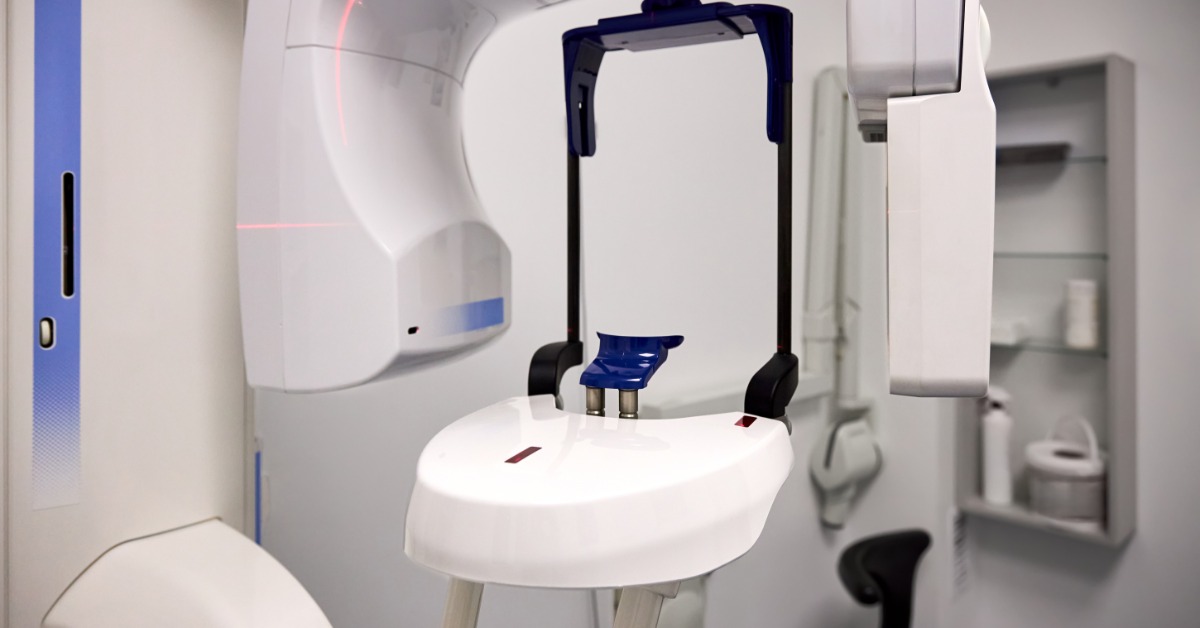 A white panoramic dental X-ray machine with blue supports and red guide lights inside a clean exam room.